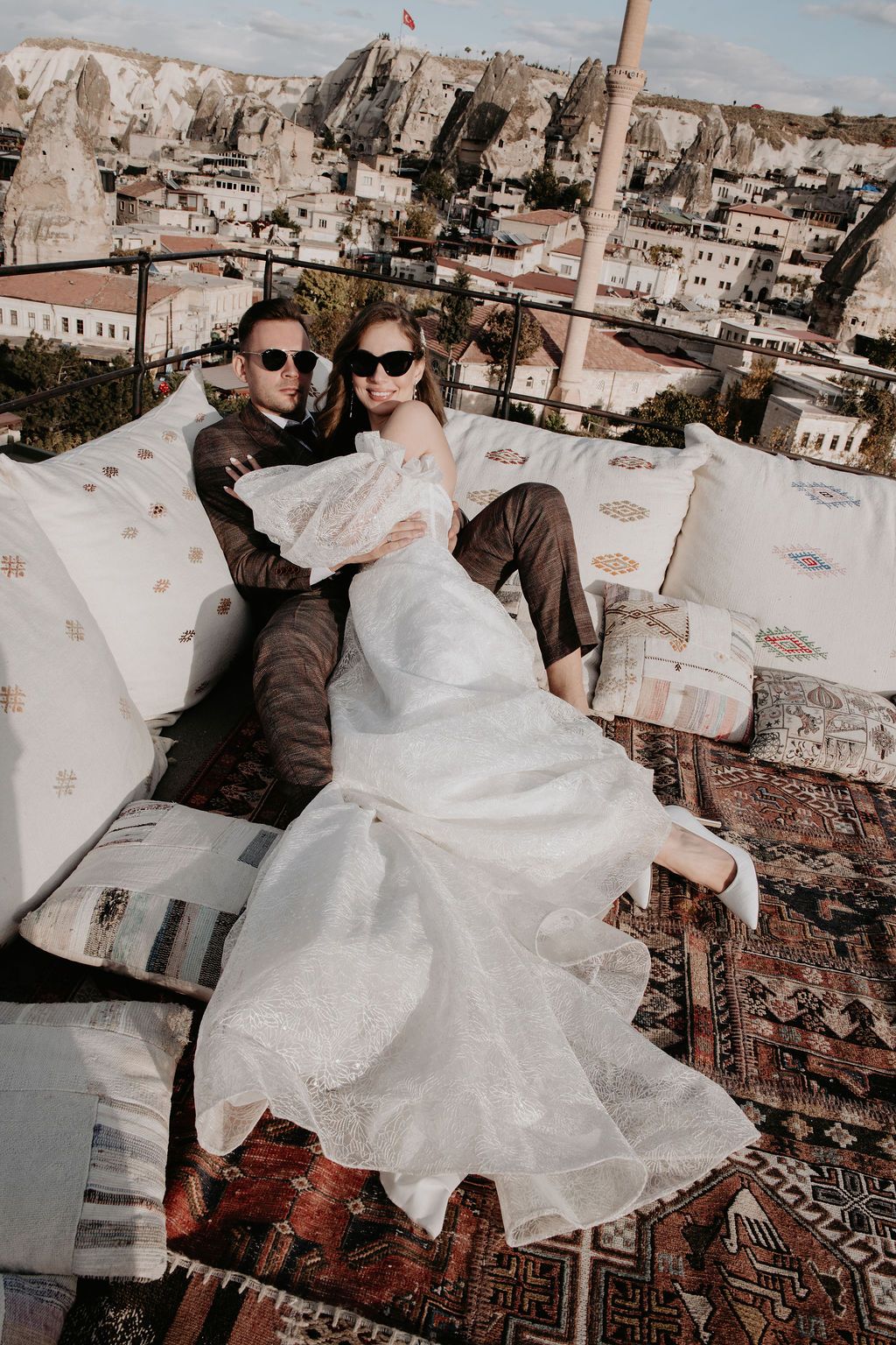 Bride and groom in sunglasses lounging on rooftop cushions with Cappadocia cave city skyline backdrop.