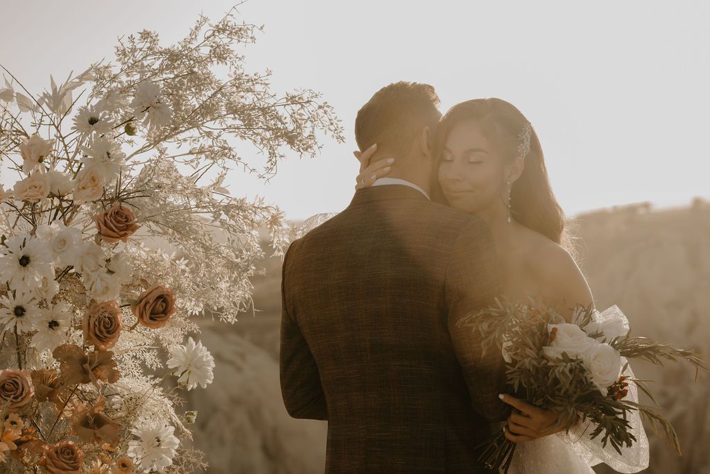 Couple embracing at golden hour wedding with boho floral arrangement of white and rust blooms.