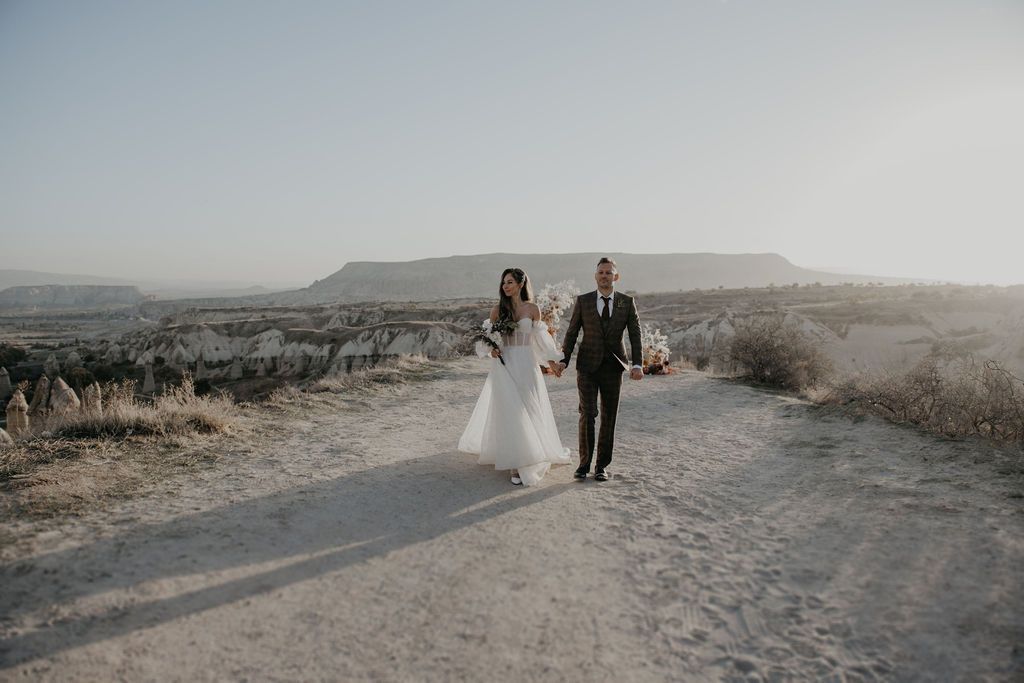 Bride and groom walking hand in hand on a desert dirt path with mesa landscape at golden hour.