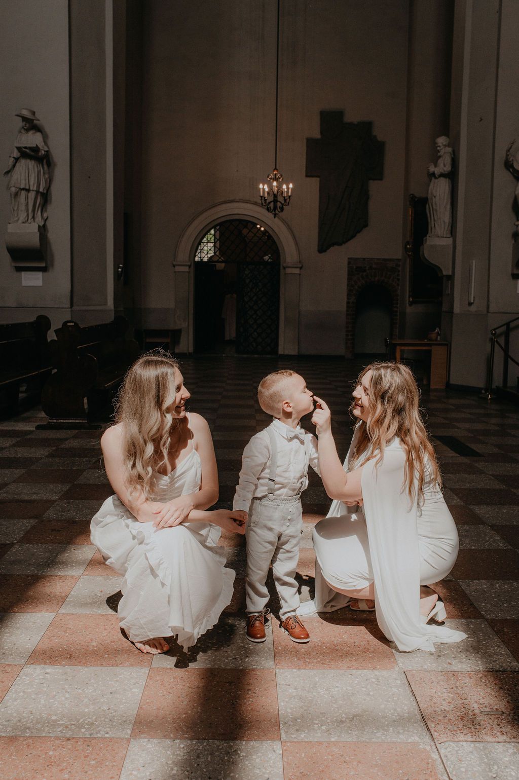 Two brides in white dresses kneel beside a young boy in a church during a wedding ceremony.