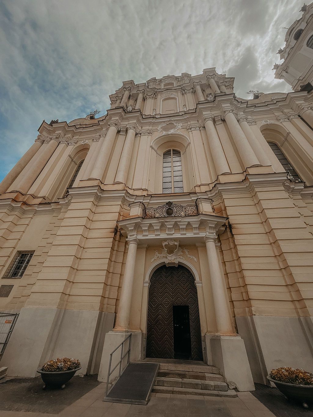 Grand baroque cathedral exterior featuring elaborate stone columns, arched doorway, and ornamental details against cloudy sky.
