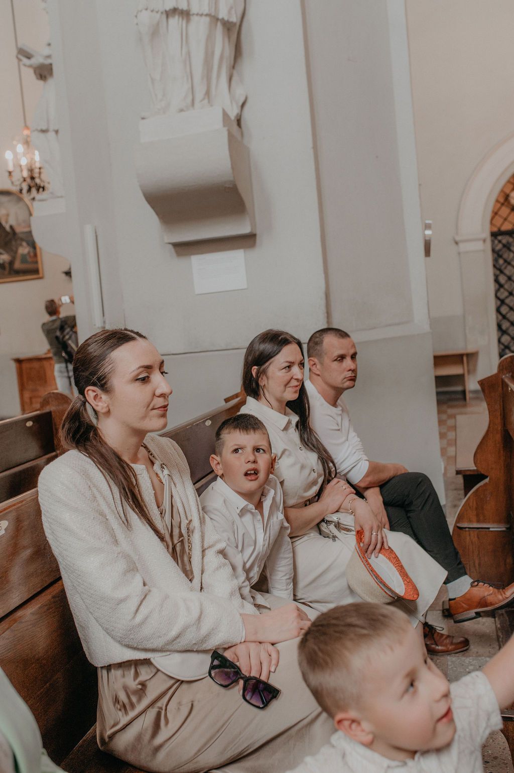 Family seated in church pews during a religious ceremony, with two young boys in white outfits.