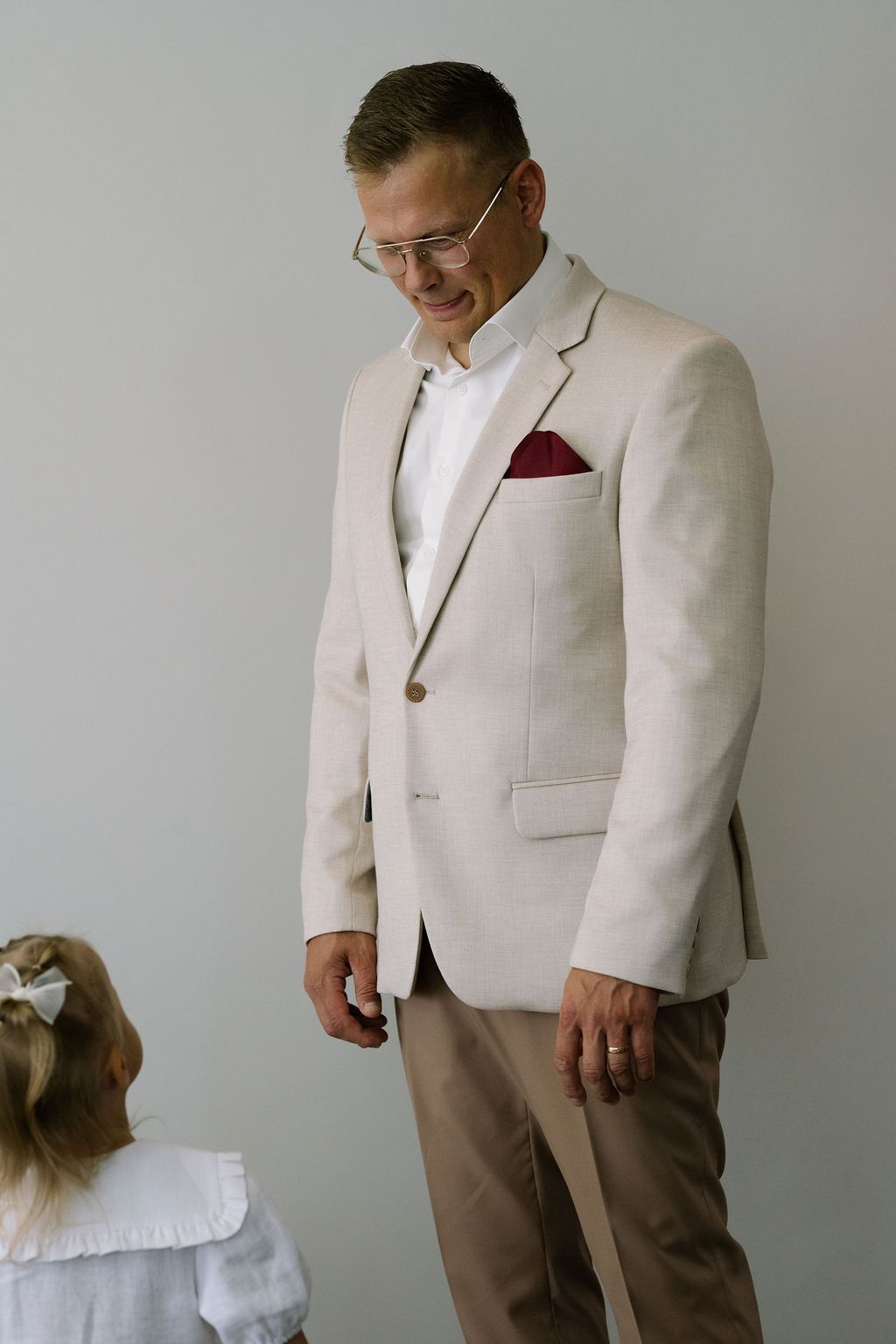 A smiling man in a beige blazer with a red pocket square looks down at a young girl with a white bow.