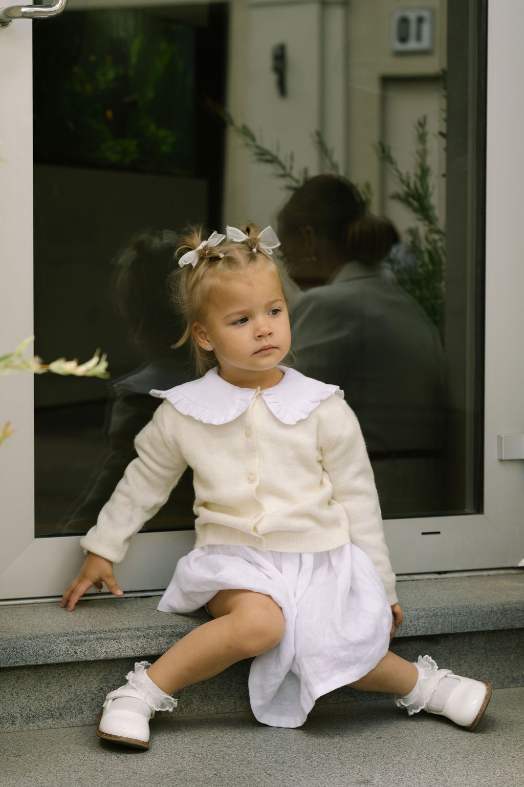 Young toddler girl in white dress and cream cardigan with bow hair clips sits on steps by glass door.