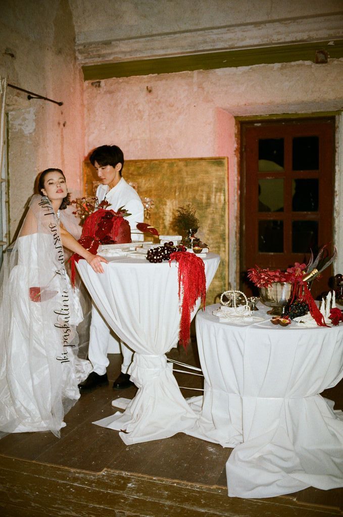 A bride in white gown arranges red floral centerpieces on draped tables in a rustic venue with peeling pink walls.