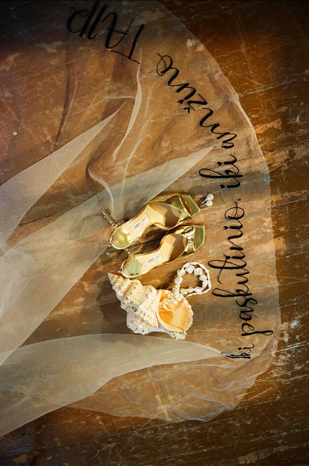 Gold bridal heels and seashell with pearl jewelry displayed on wood surface under flowing white veil with cursive script.