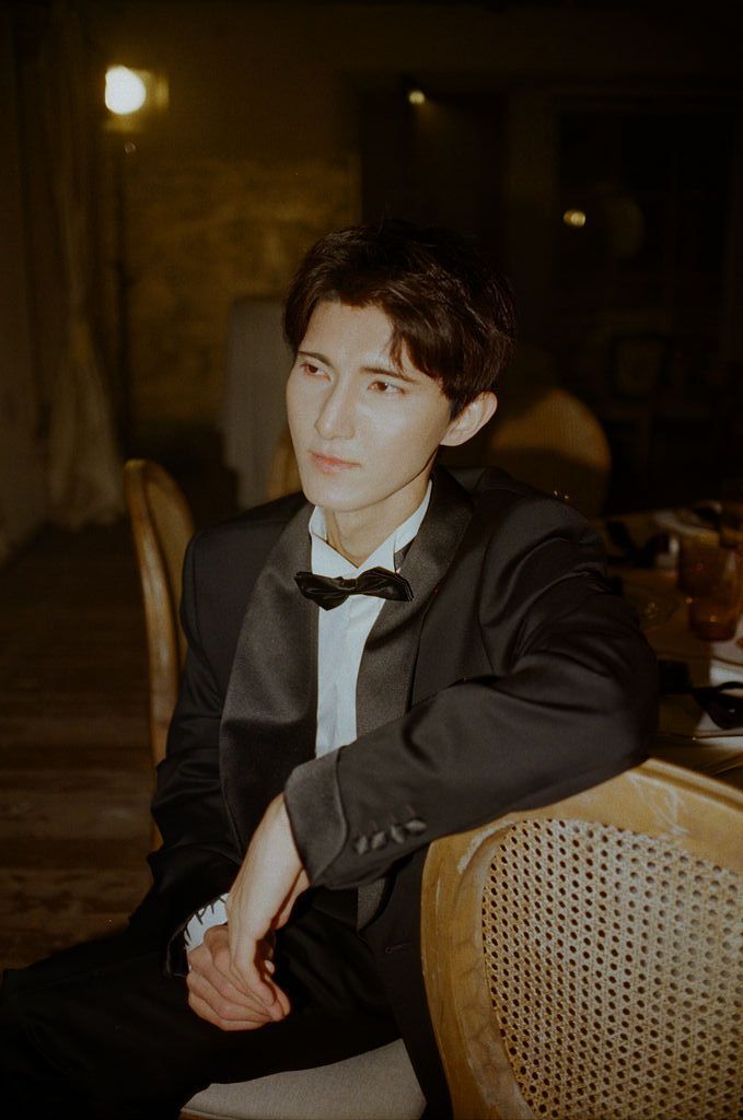 Young man in black tuxedo and bow tie seated elegantly in a cane chair under warm ambient lighting.