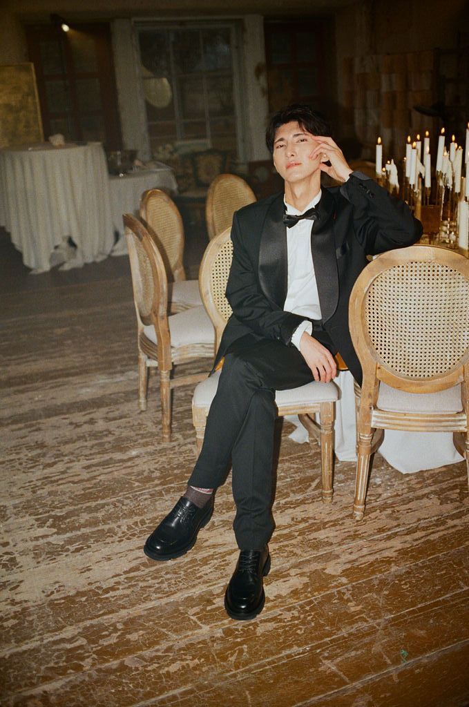 Young man in black tuxedo with bow tie seated elegantly on ornate chair in candlelit formal venue.