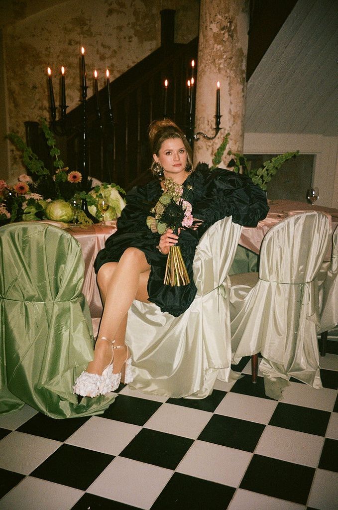 Woman in black dress holding bouquet sits on satin chair at elegant event with checkered floor and candlelight.