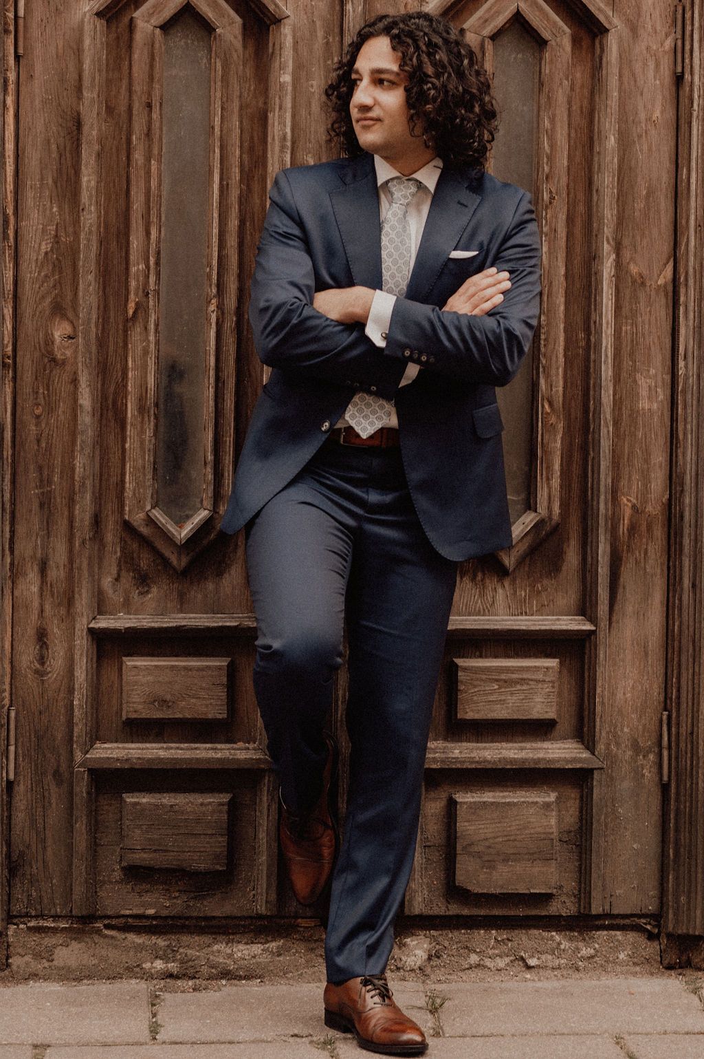A young man with curly hair wearing a navy blue suit, patterned tie, and brown oxford shoes, posing confidently against a wooden door.