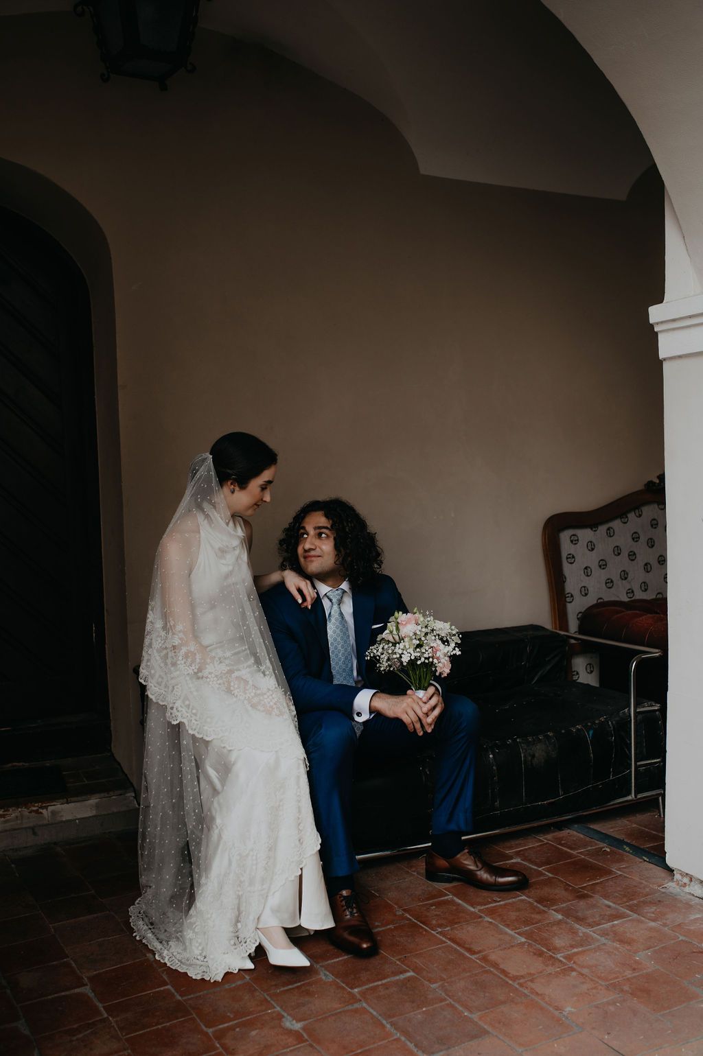 Bride in lace gown and veil stands beside groom in navy suit holding bouquet, seated on vintage sofa indoors.