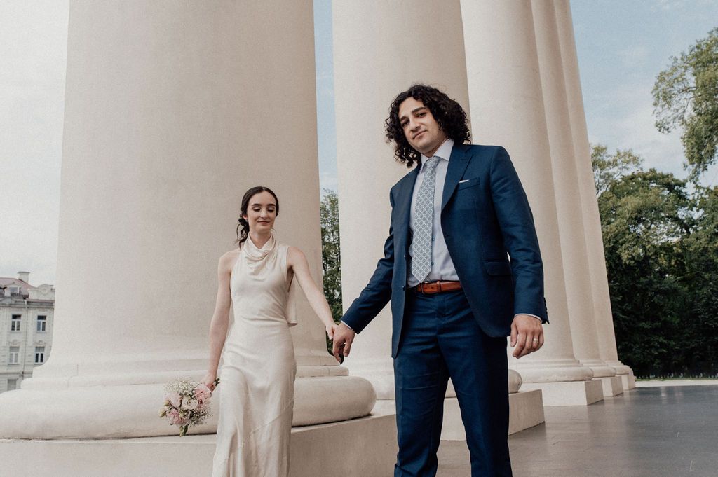 A bride and groom hold hands at a columned venue, she in a white gown, he in a navy suit.