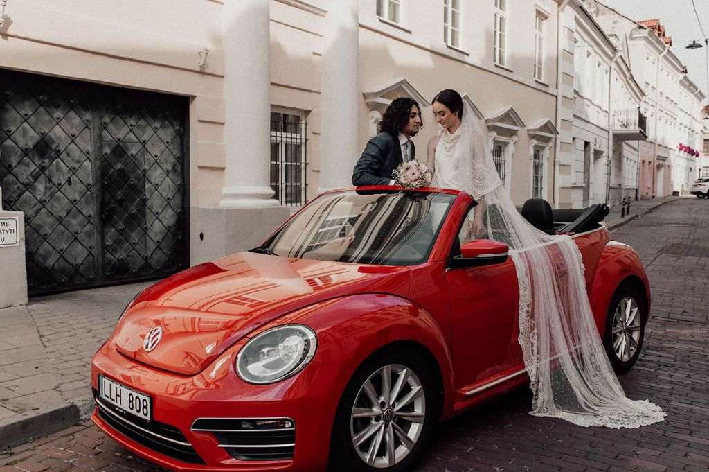 Two brides posing on a red Volkswagen Beetle convertible on a charming European cobblestone street.