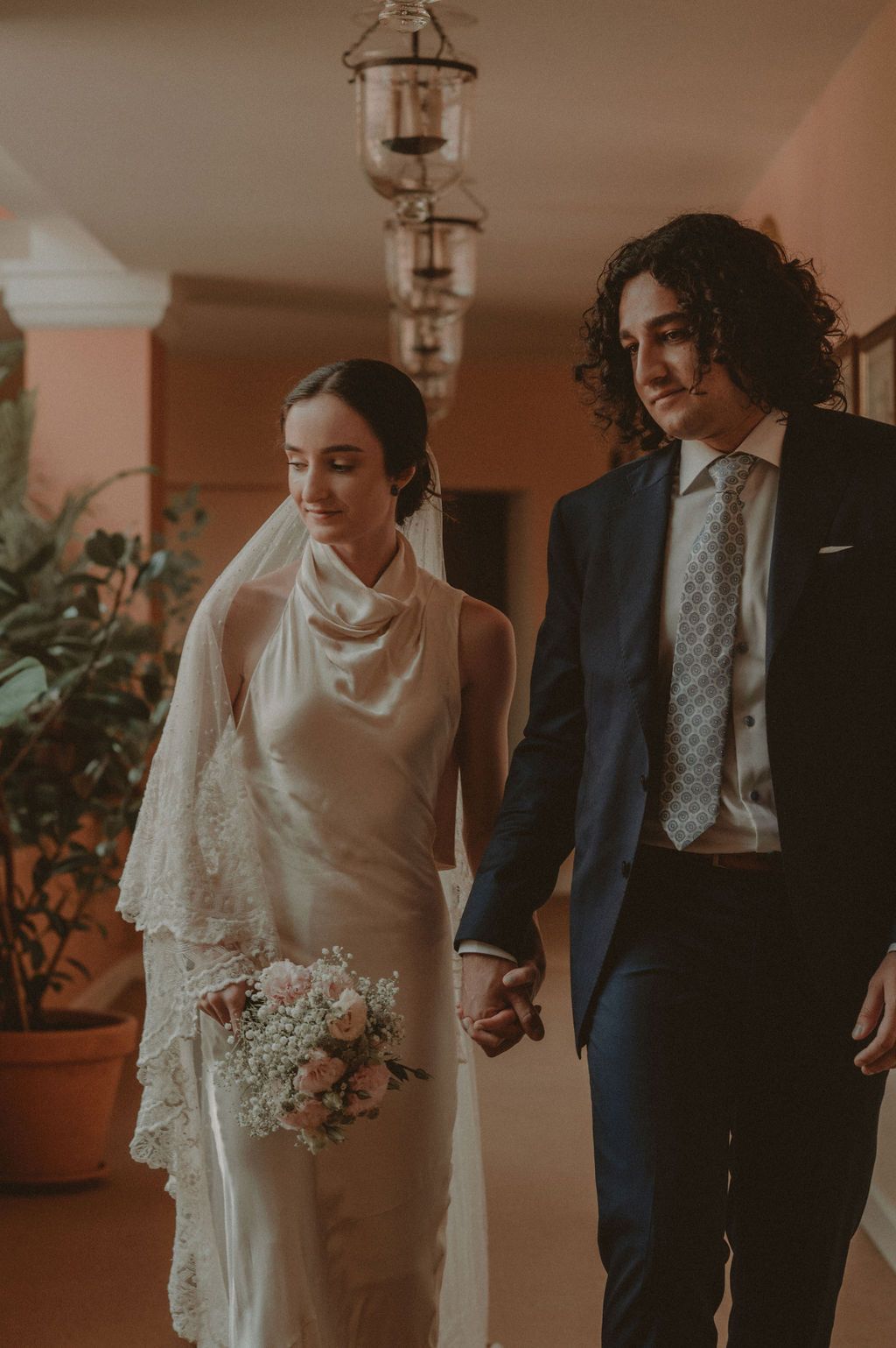 A bride in white gown holding bouquet walks hand-in-hand with groom in navy suit under chandelier.