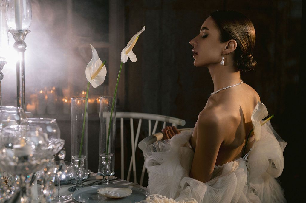 Elegant bride in backless white gown admires calla lily at candlelit table with crystal glassware.