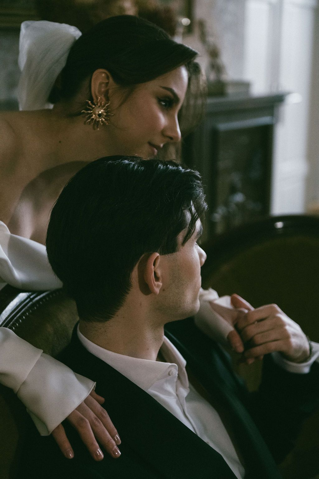 Elegant bride in white gown with gold star earring embraces groom in black tuxedo in a moody vintage interior.