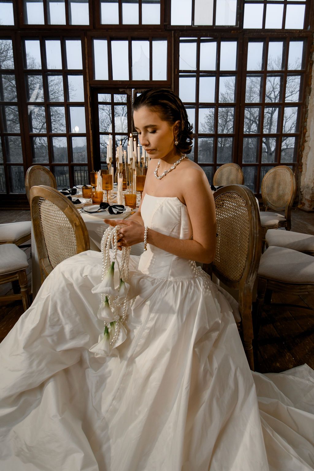 Bride in white strapless gown seated at elegant reception table, holding pearl bouquet, with candles and bottles in background.