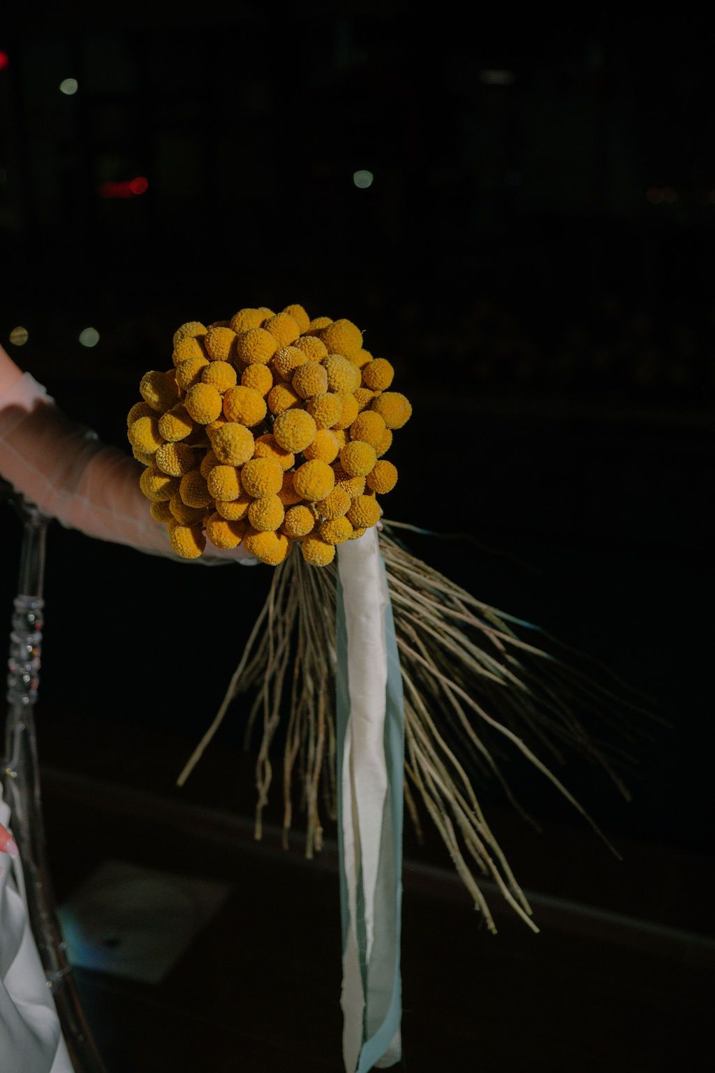 Hand holding a vibrant yellow craspedia billy ball flower bouquet with ribbon streamers against dark background.