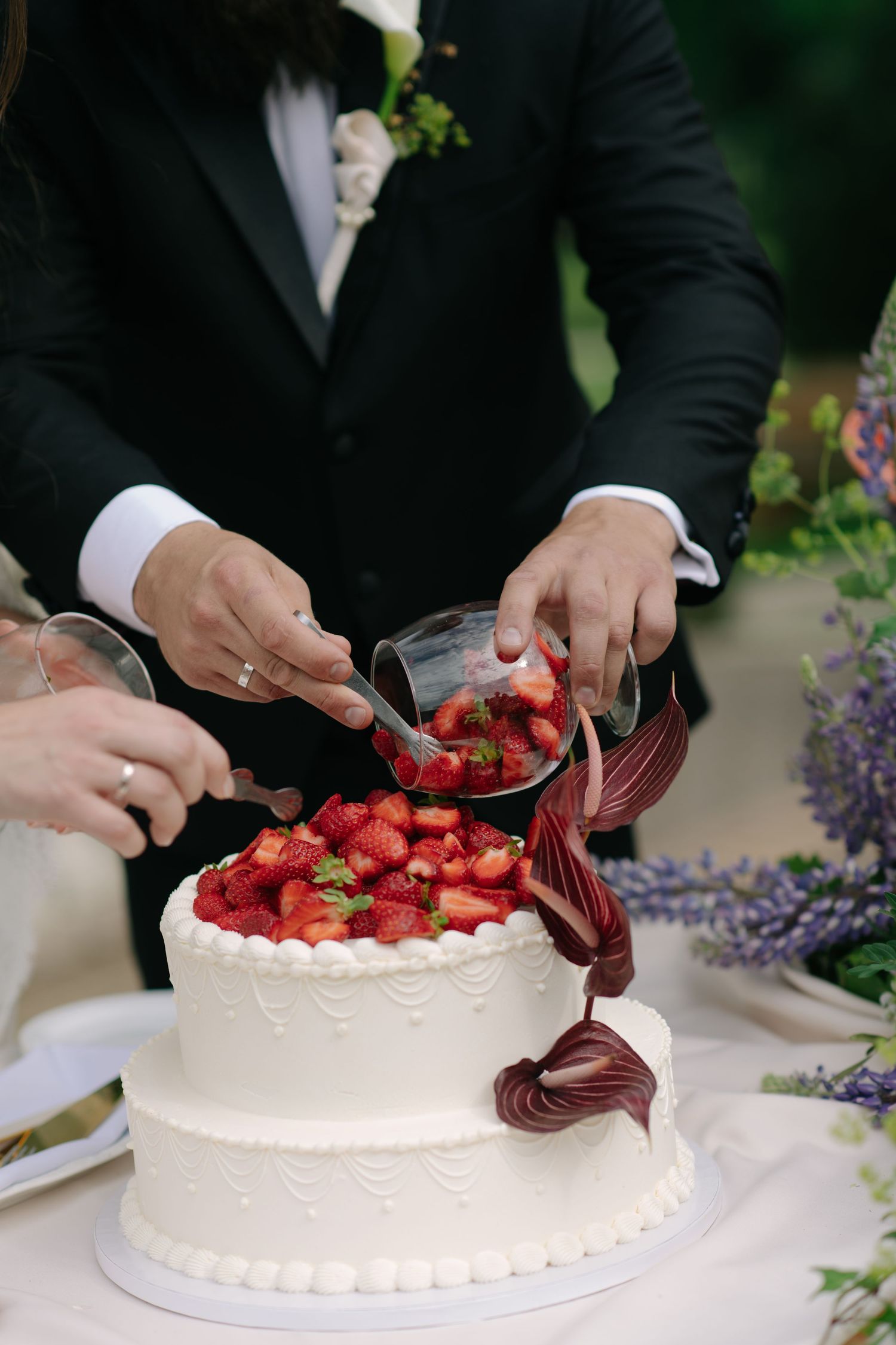 Newlyweds cut two-tier white wedding cake topped with fresh strawberries and calla lily decoration outdoors.