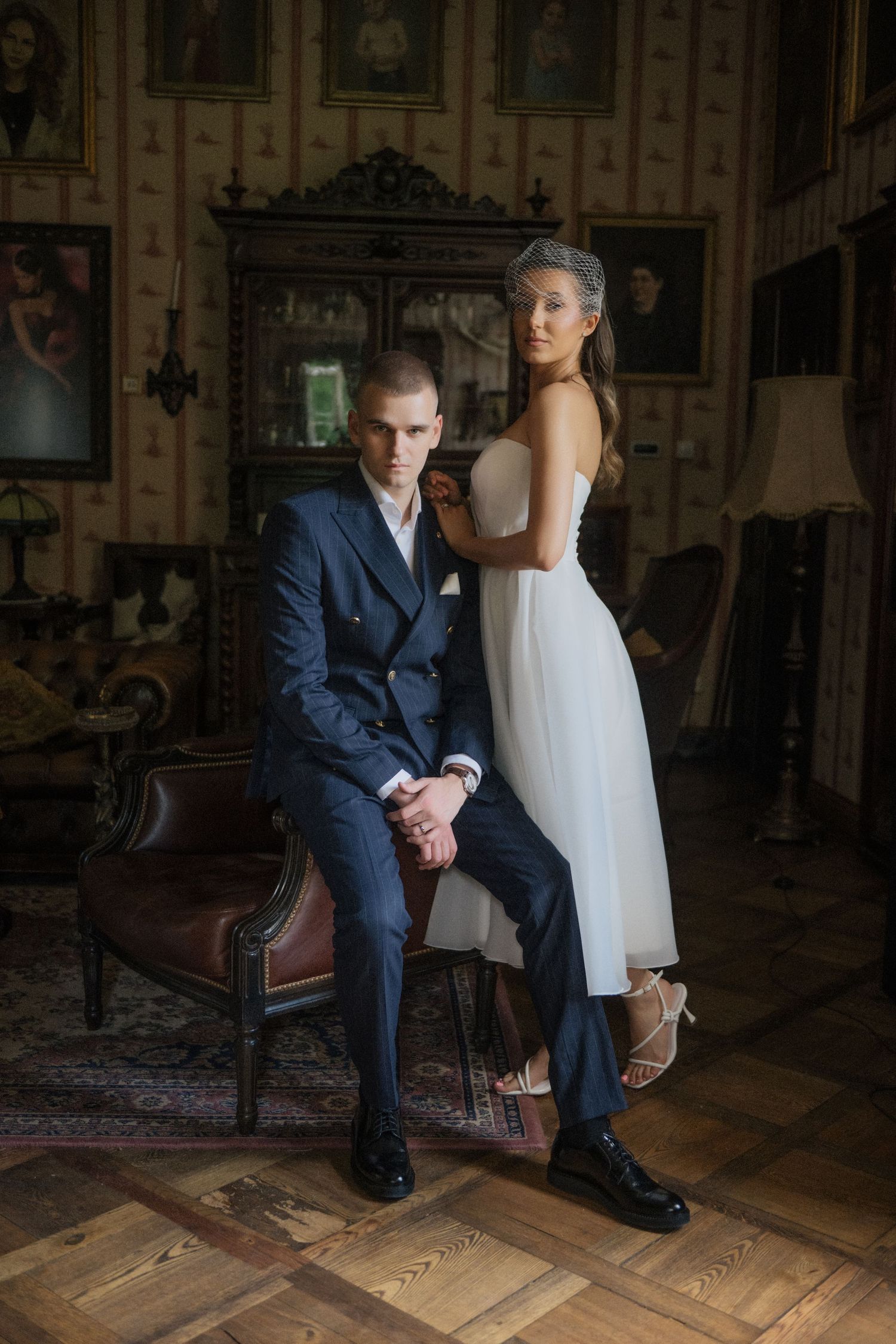 Elegant wedding couple posing in vintage room, groom in navy suit seated, bride in white dress with veil standing beside him.