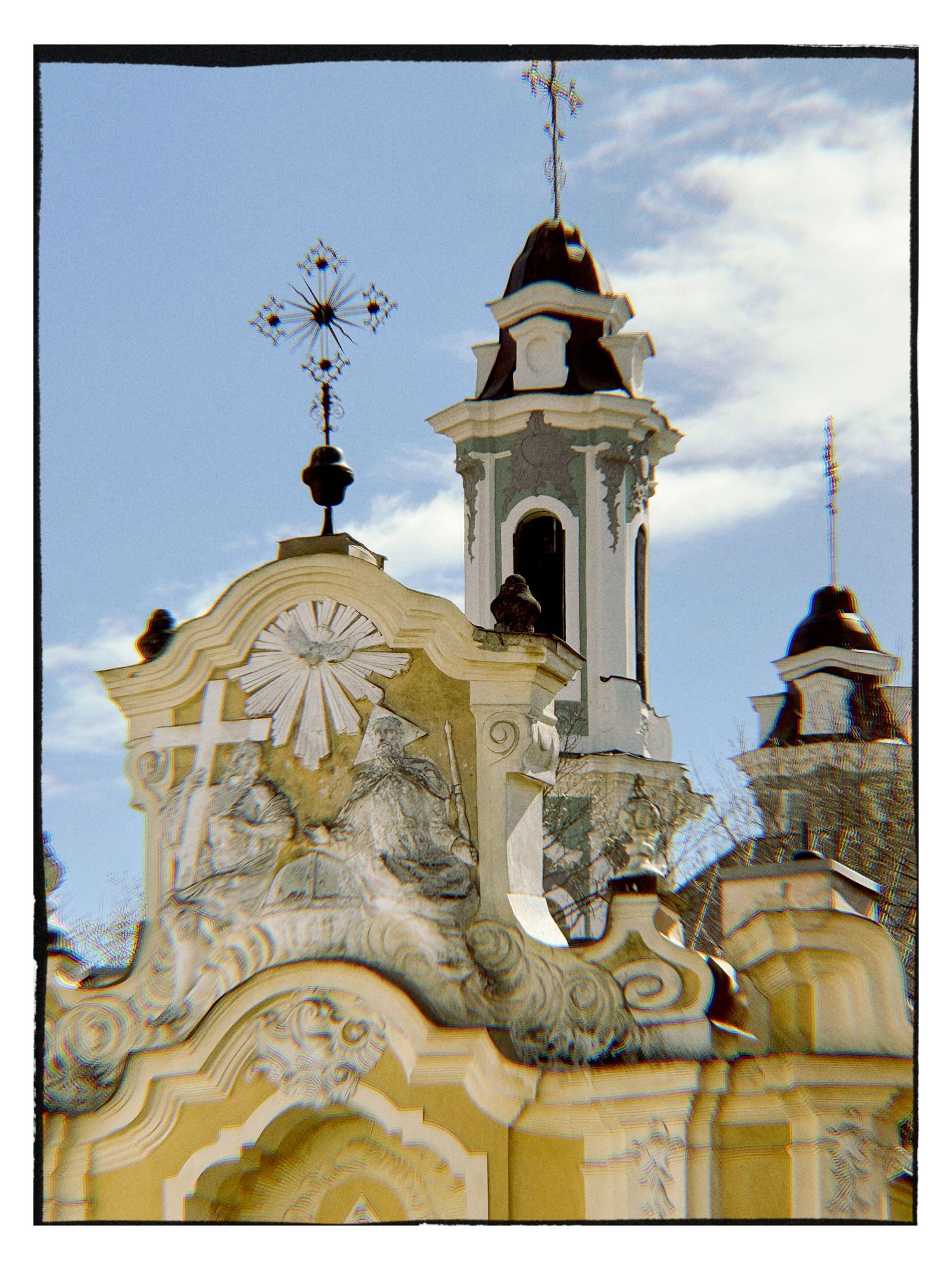 Ornate yellow Baroque church facade with decorative crosses, bell tower, and intricate stone carvings against a blue sky.