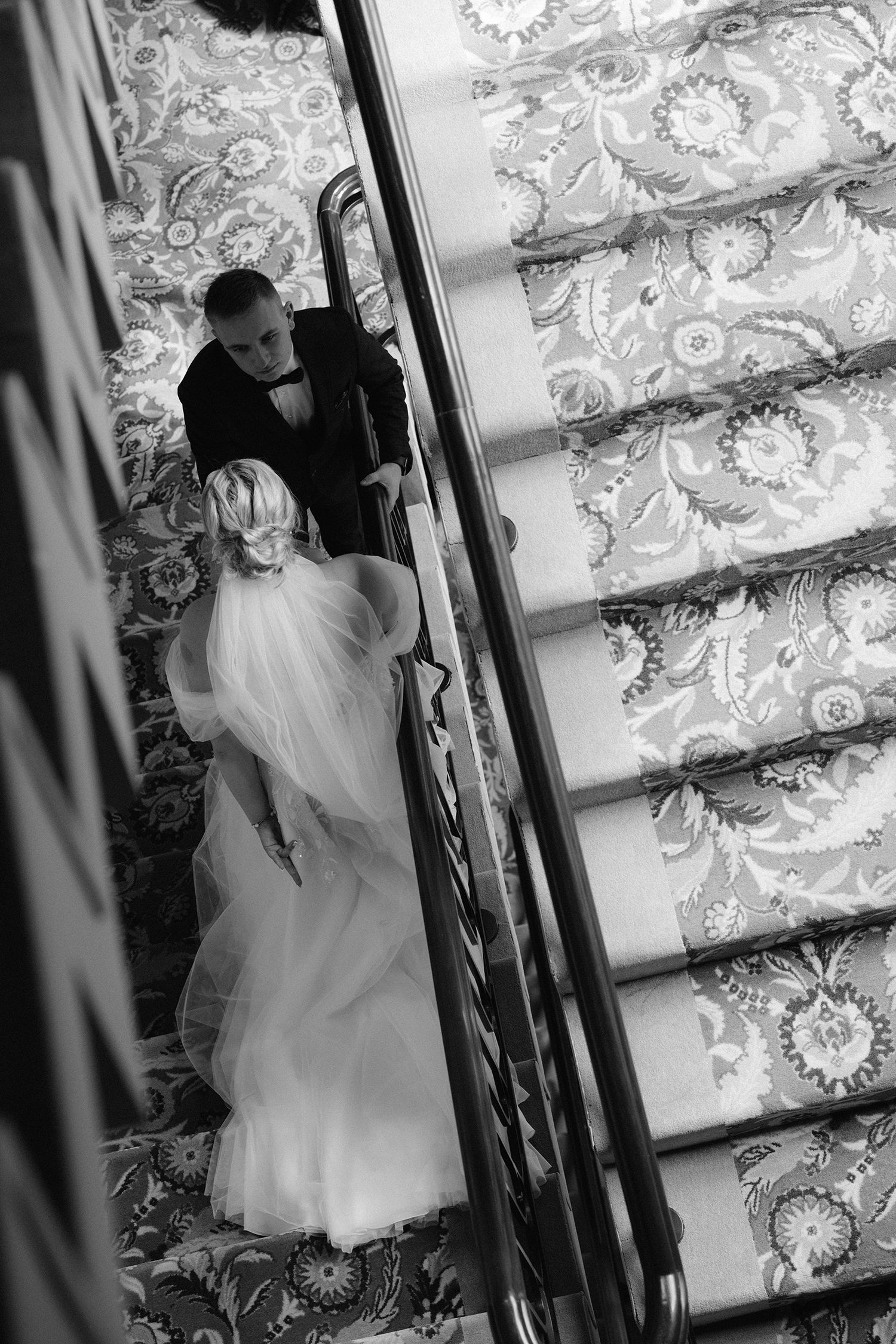 A bride in a white gown descends an ornate staircase with floral wallpaper in a black and white photograph.