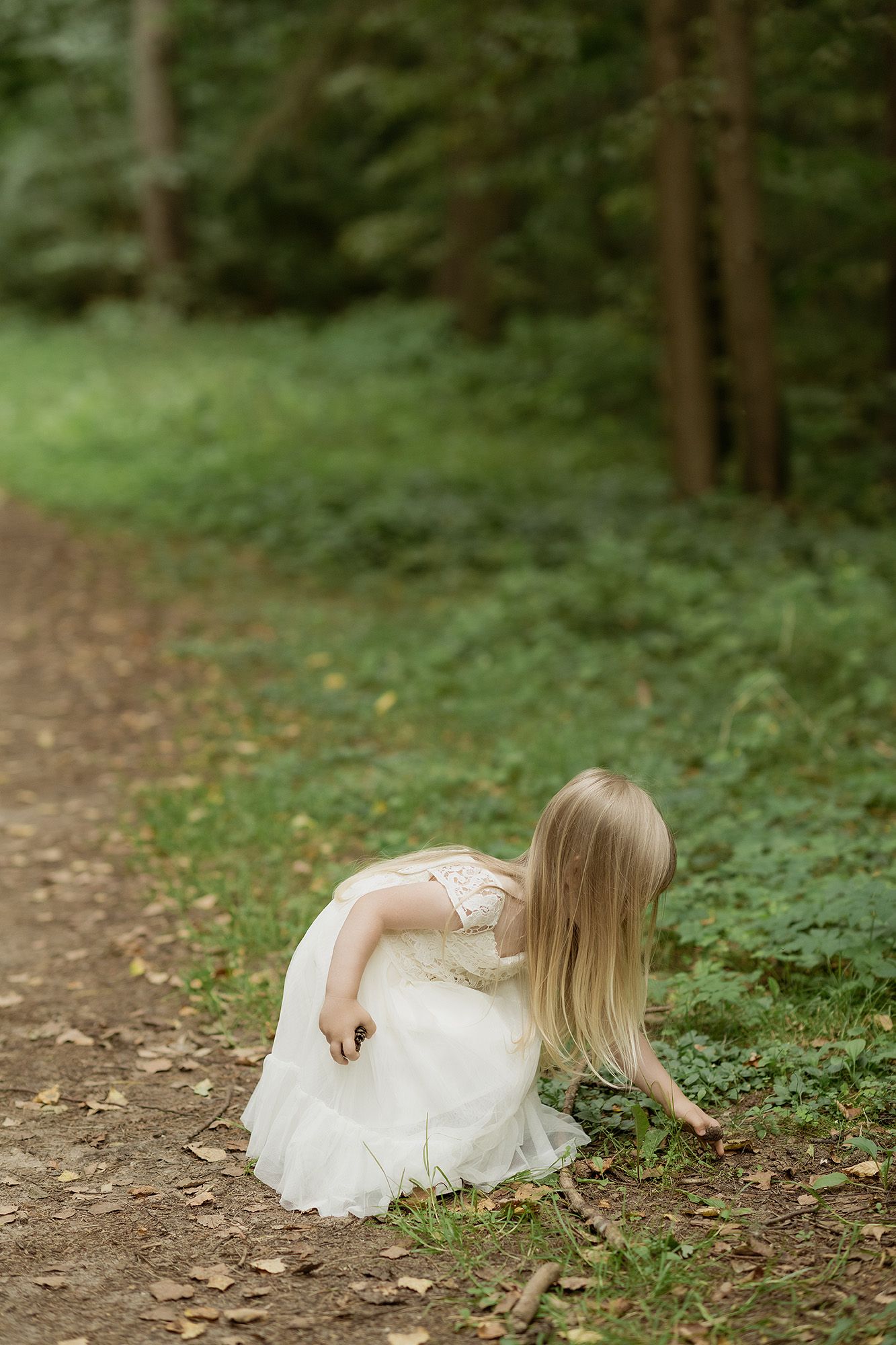 A person in a white dress crouches on a dirt path in a lush green forest setting.