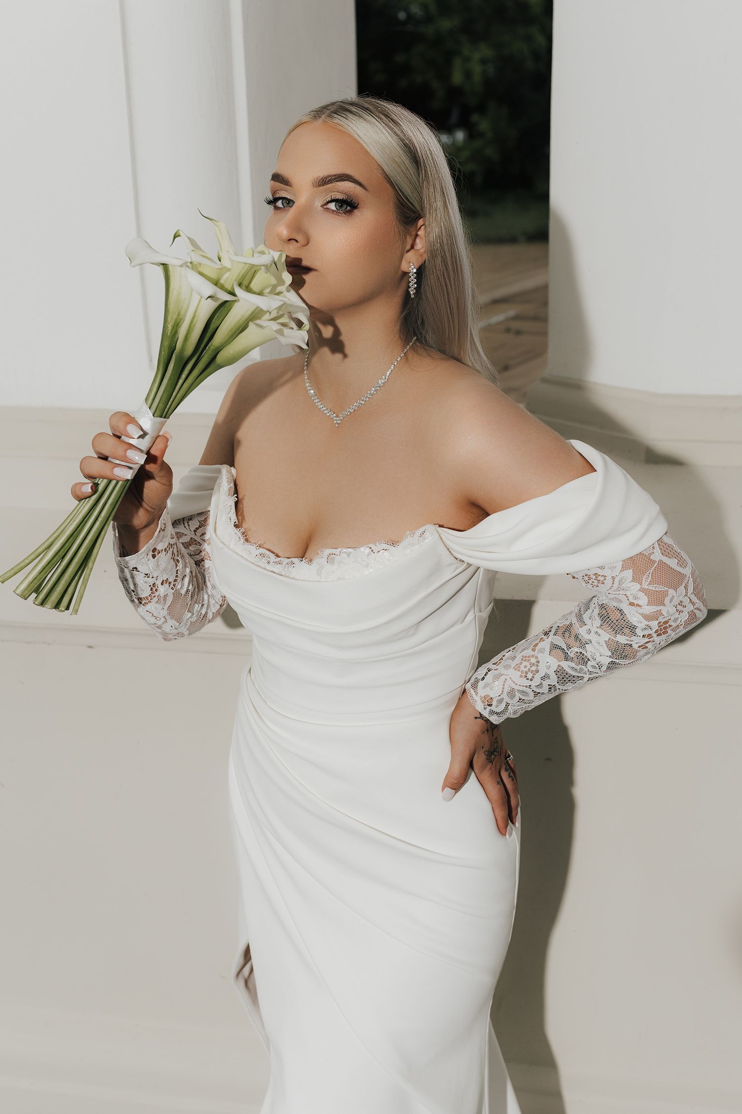 A bride in an off-shoulder white wedding gown with lace sleeves poses elegantly against a light backdrop.