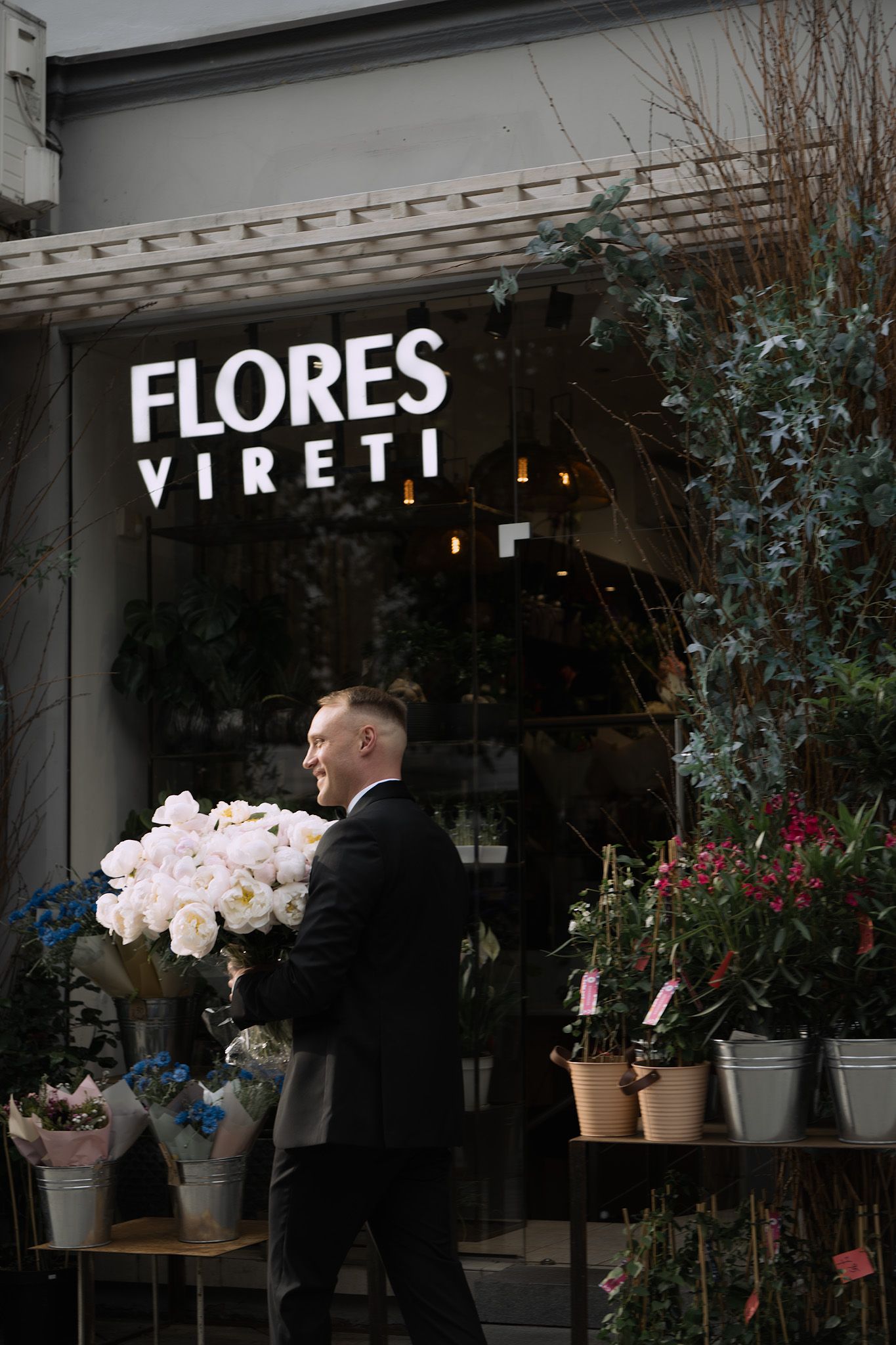 A person carries a large bouquet of pink flowers outside the Flores Vireti flower shop storefront.