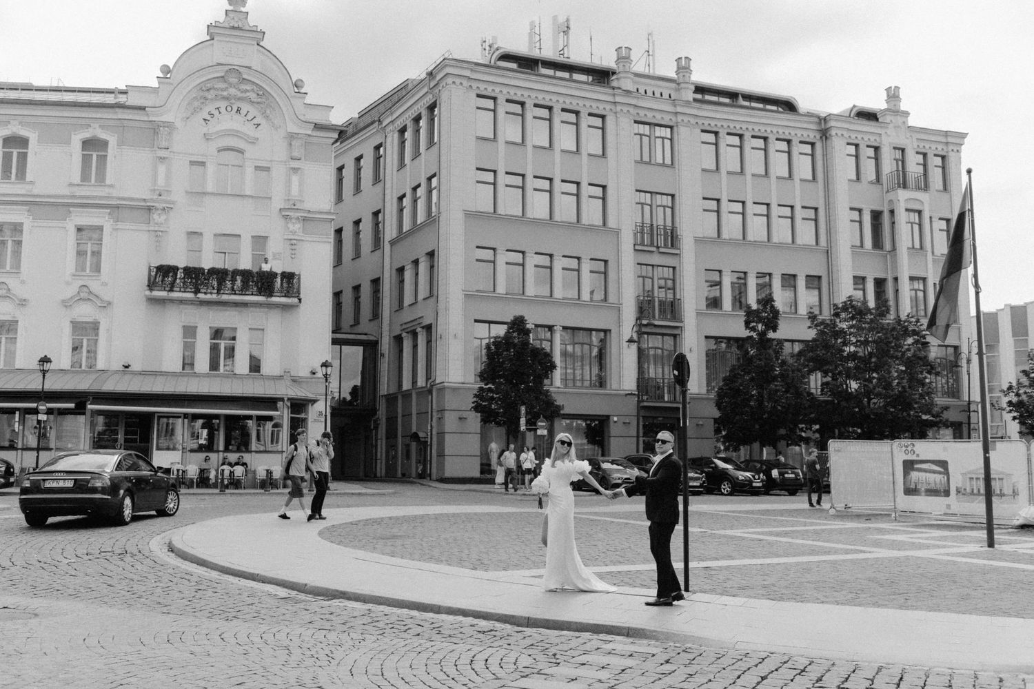 Black and white photo of a bride and groom posing on a city street corner with historic buildings, trees, and parked cars behind them.