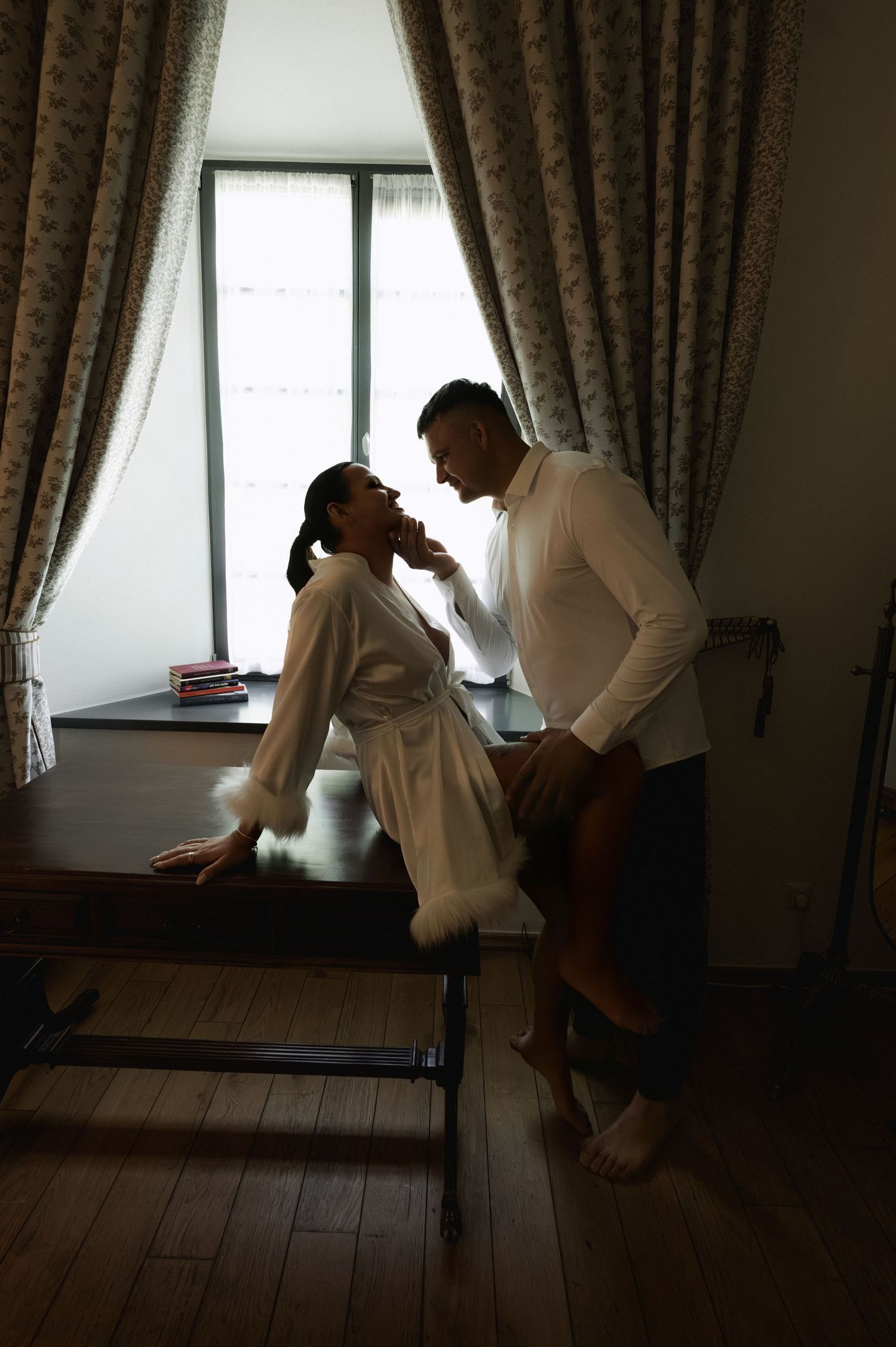 Silhouetted couple in a moody, softly lit room, a woman in a feathered-trim robe sitting on a wooden table beside a man in a white shirt.