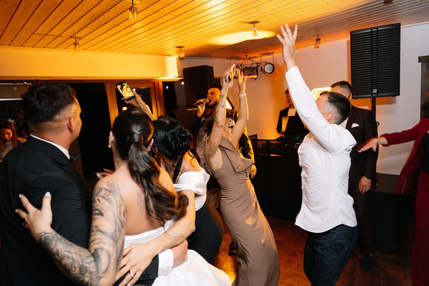Wedding guests dance and cheer with raised arms at an indoor reception while a speaker and warm ceiling lights glow in the background.