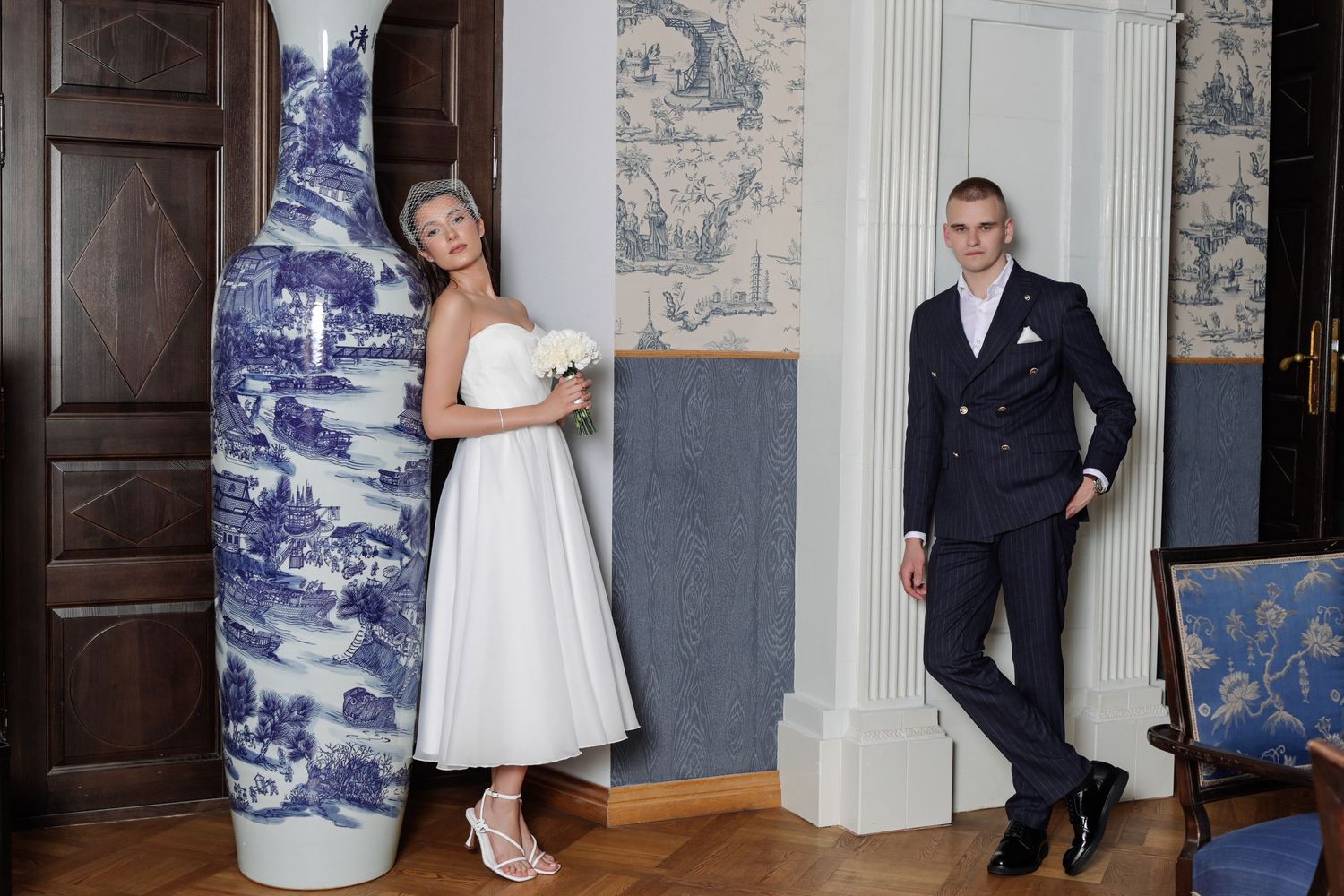 Bride in a white strapless dress holds a bouquet beside a large blue and white vase while the groom in a navy suit leans against a column.