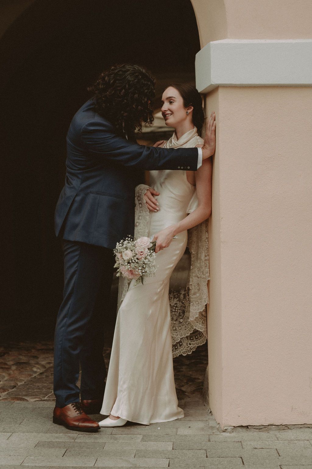 Bride in white gown holds bouquet as groom in navy suit leans against pink wall during romantic wedding portrait.