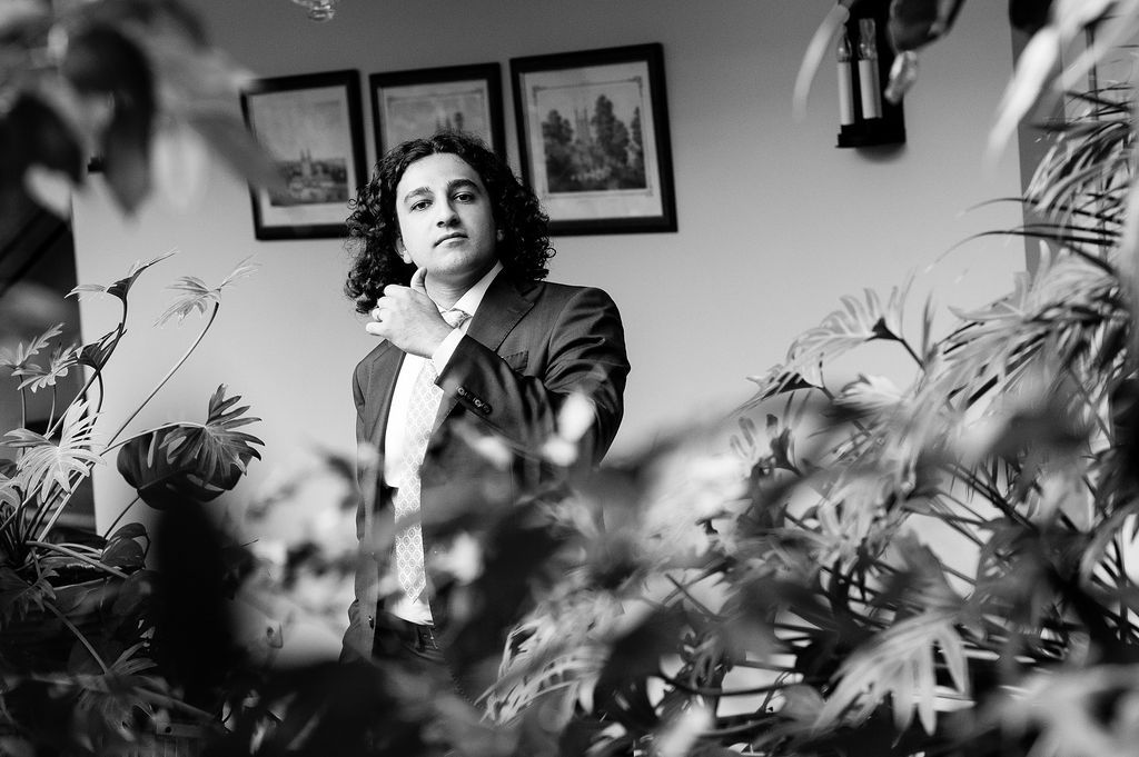 Black and white portrait of a person among lush indoor plants with framed artwork on the wall behind.