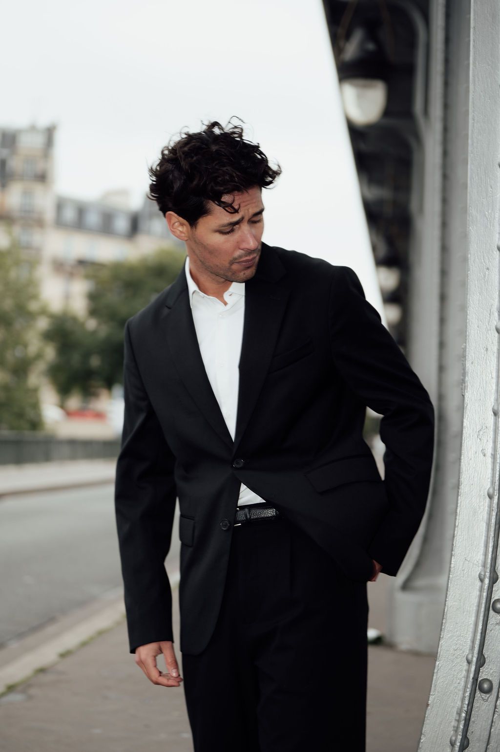 A stylish man in a black suit and white shirt poses outdoors on a city street with curly dark hair.