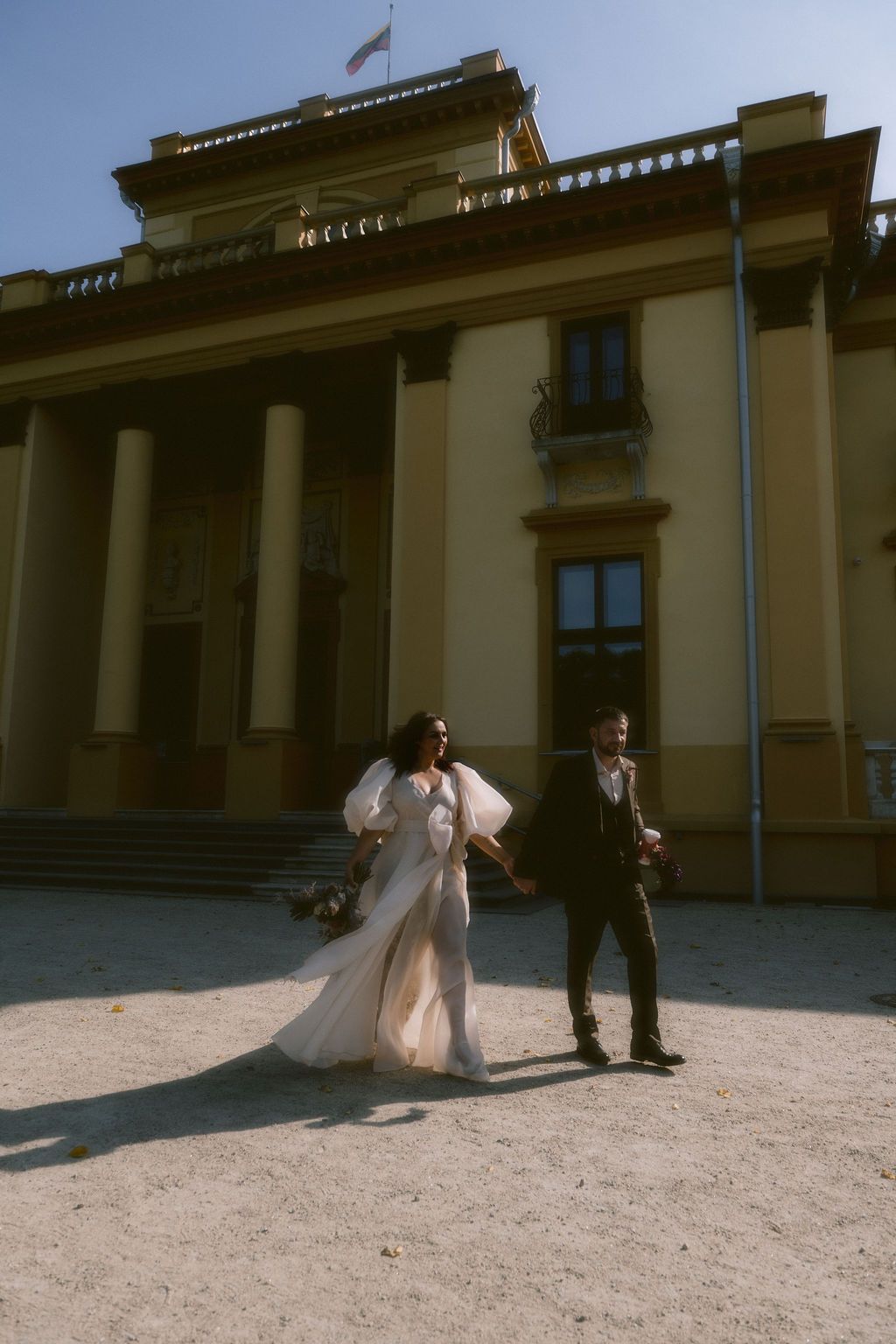 Bride in white gown and groom in dark suit walk hand in hand before a grand yellow neoclassical building.