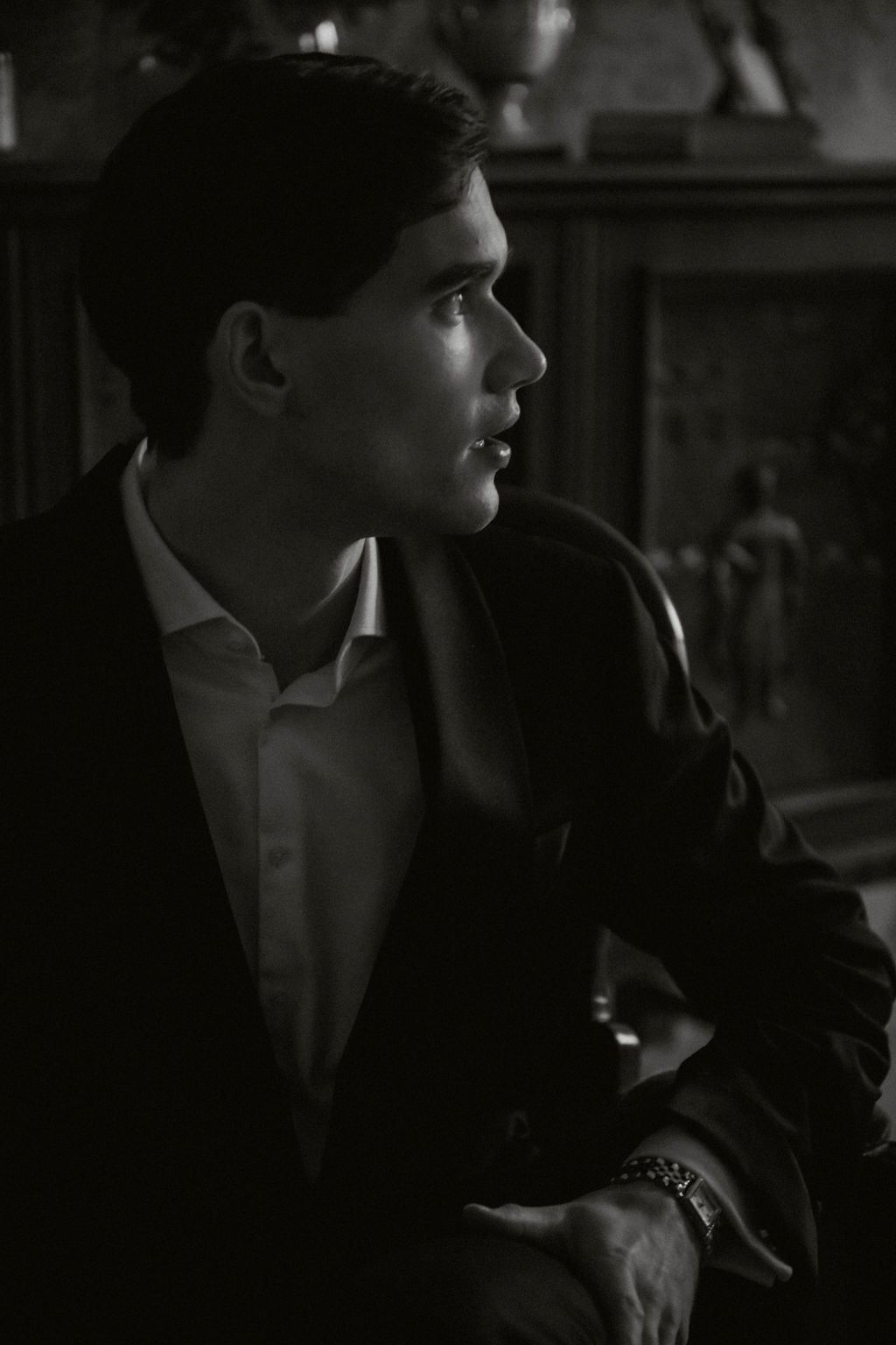 Elegant young man in black suit captured in dramatic black and white profile portrait indoors.