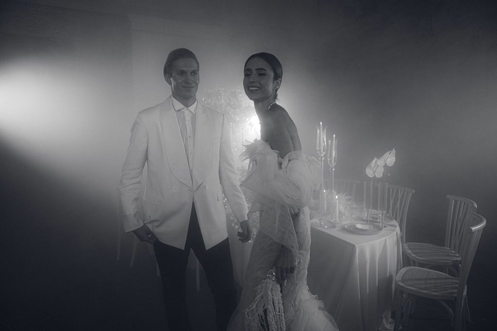 Elegant couple in black and white photo, groom in white tuxedo and bride in feathered gown at wedding reception.