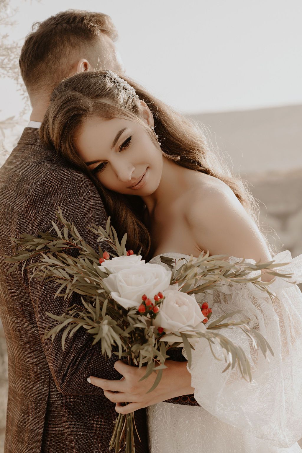 Bride in white dress hugging groom in brown suit, holding bouquet of white roses and greenery at outdoor wedding.