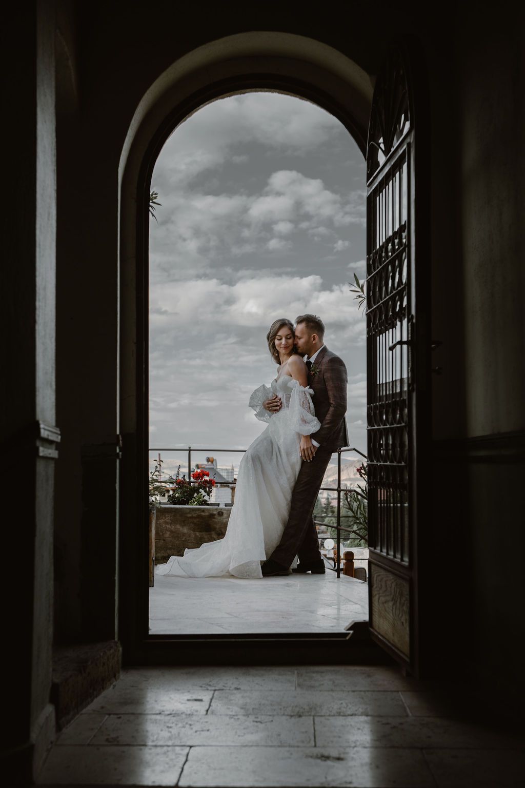 Bride and groom embracing in an arched doorway, framed by dramatic cloudy skies on a romantic outdoor balcony.