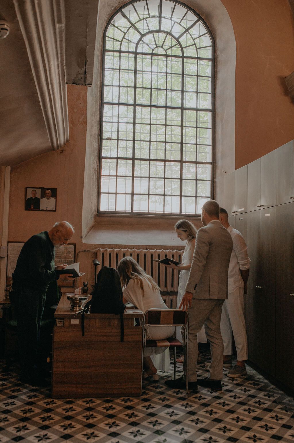 People gathered around a baptismal font in a historic church with arched stained glass windows and ornate tiled floors.