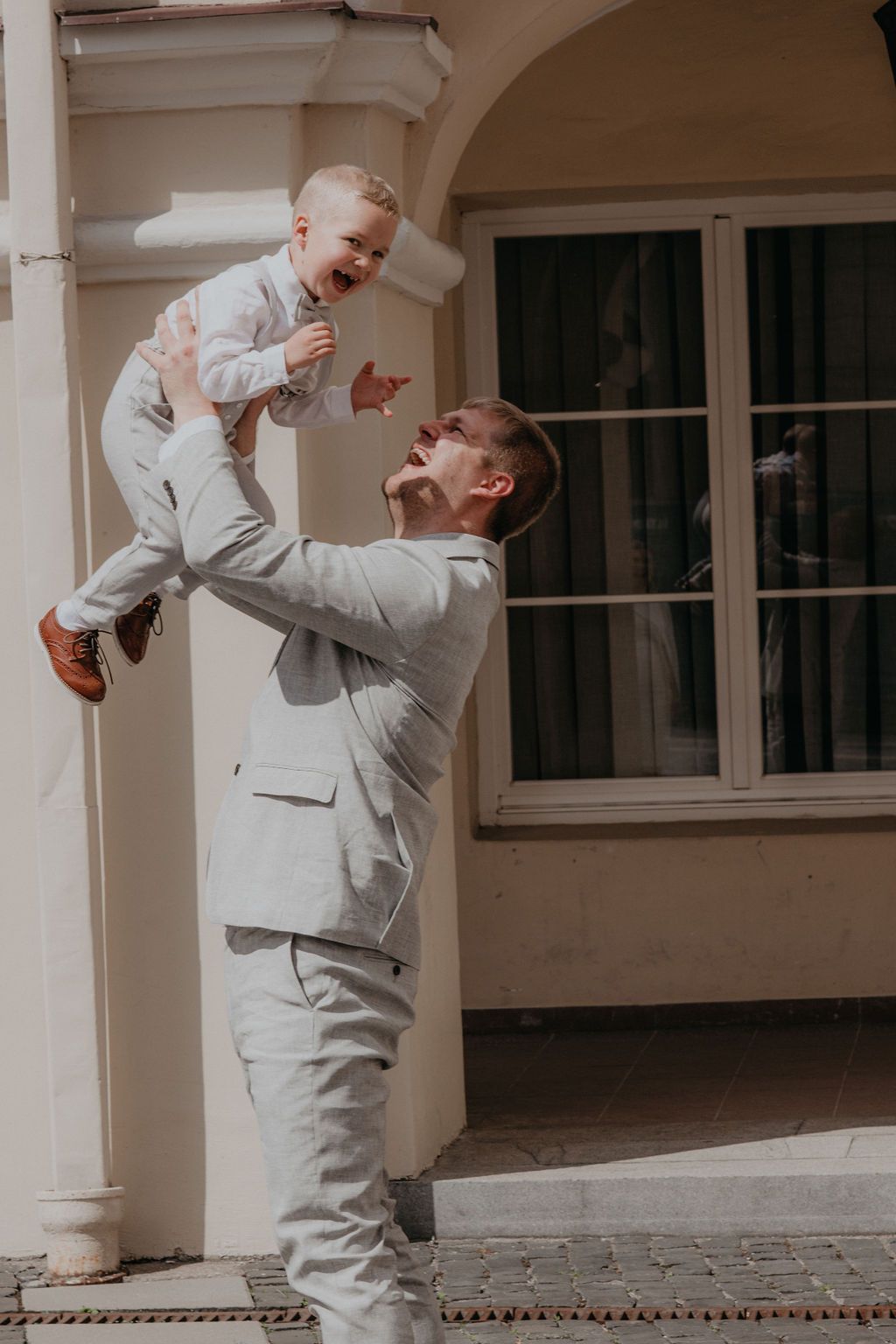 Man in light suit joyfully lifting laughing baby outdoors near elegant building arch.