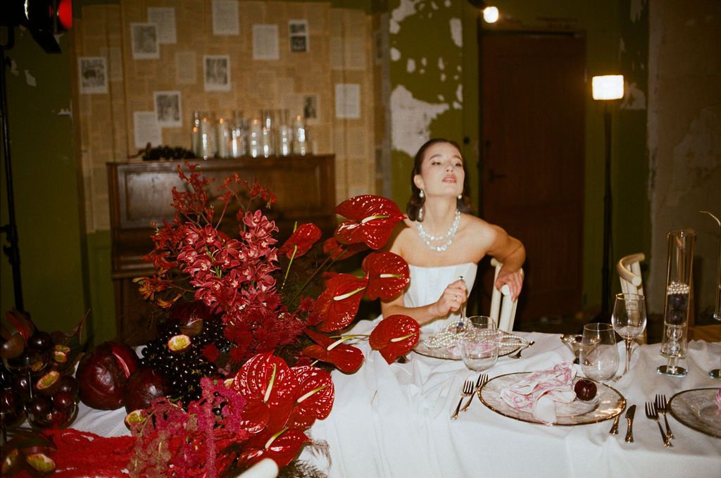 Bride in white dress sits at elegantly set table with lush red floral arrangements including anthuriums at a wedding reception.