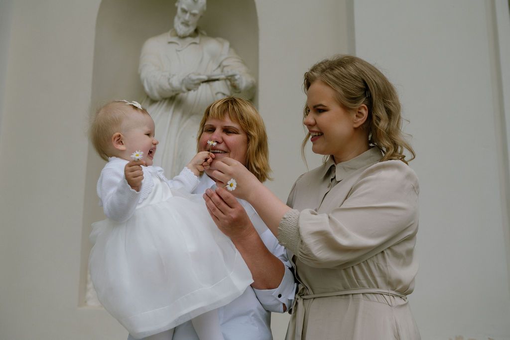 Two women and a baby in white dress celebrate a baptism ceremony near a religious statue indoors.