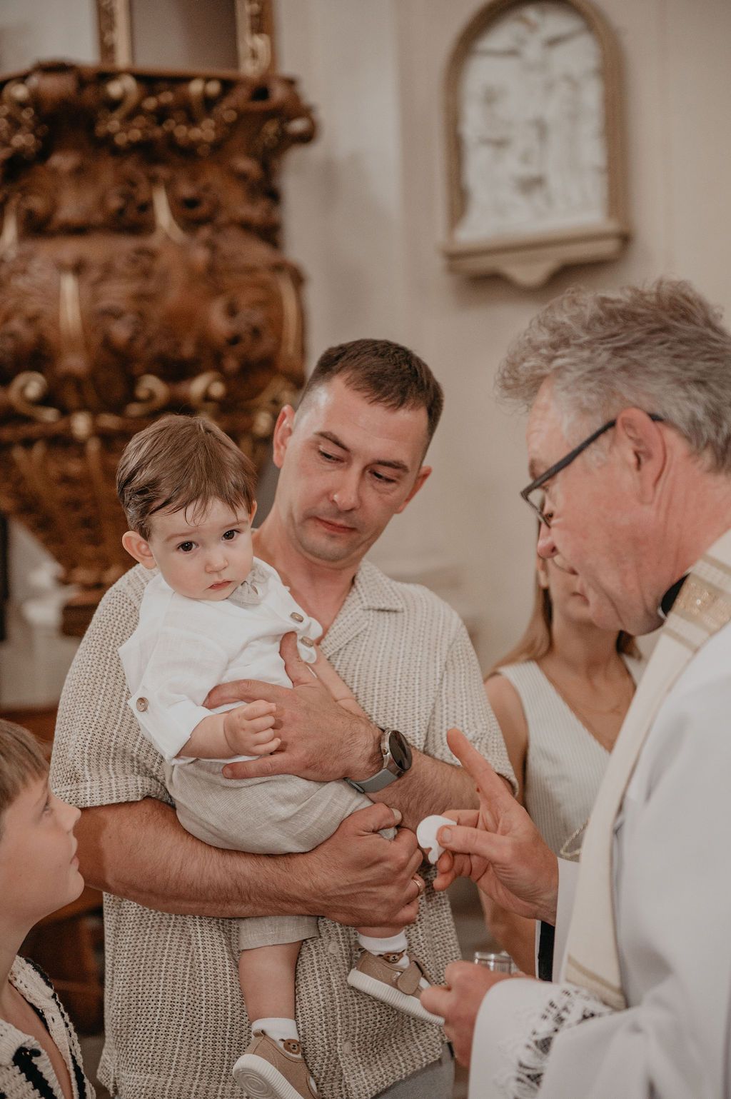 A priest performs a baptism ceremony for a baby held by his father inside an ornate church.