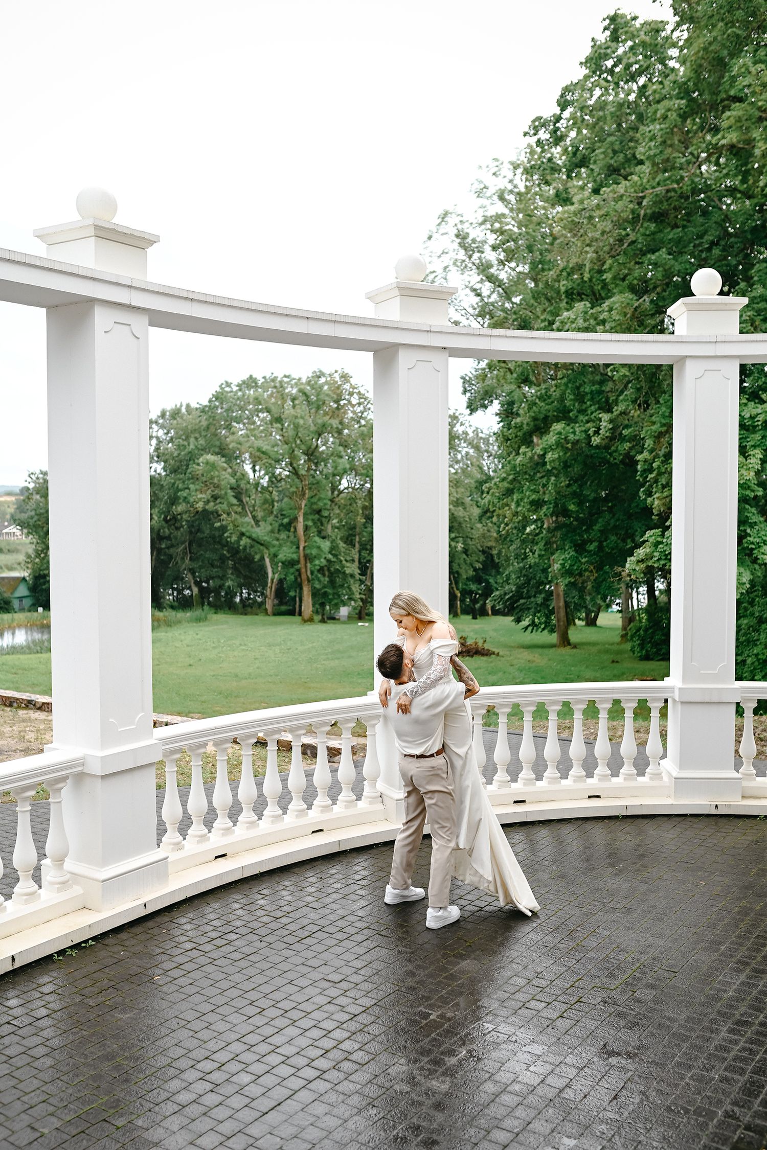 A couple in white outfits dances together on an elegant white columned porch overlooking a lush green lawn.