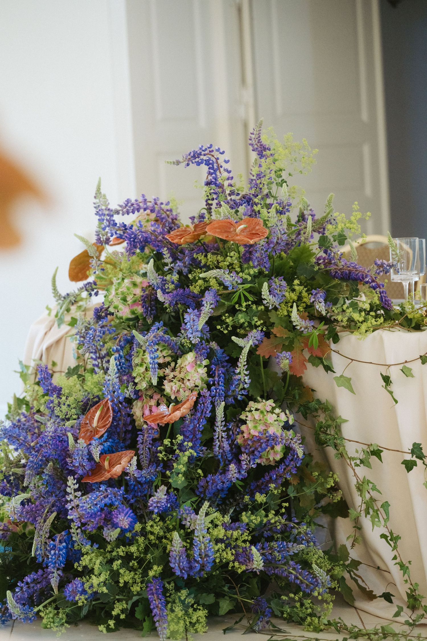 Lush wedding floral arrangement featuring blue delphiniums, orange anthuriums, and trailing ivy on a draped table.