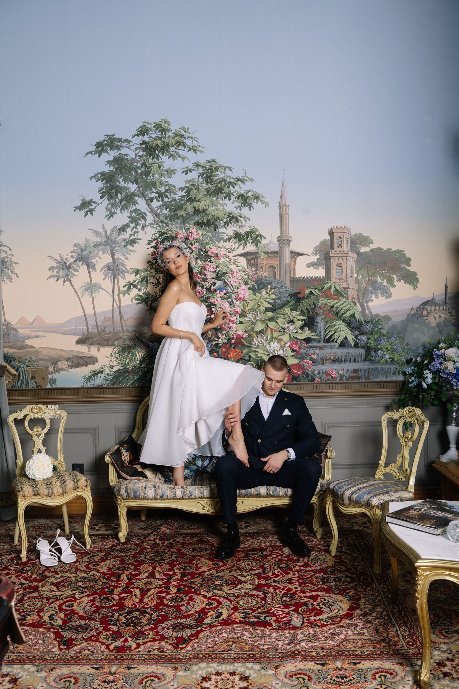 Bride in white gown poses with groom in black suit on ornate gold sofa before scenic mural backdrop.