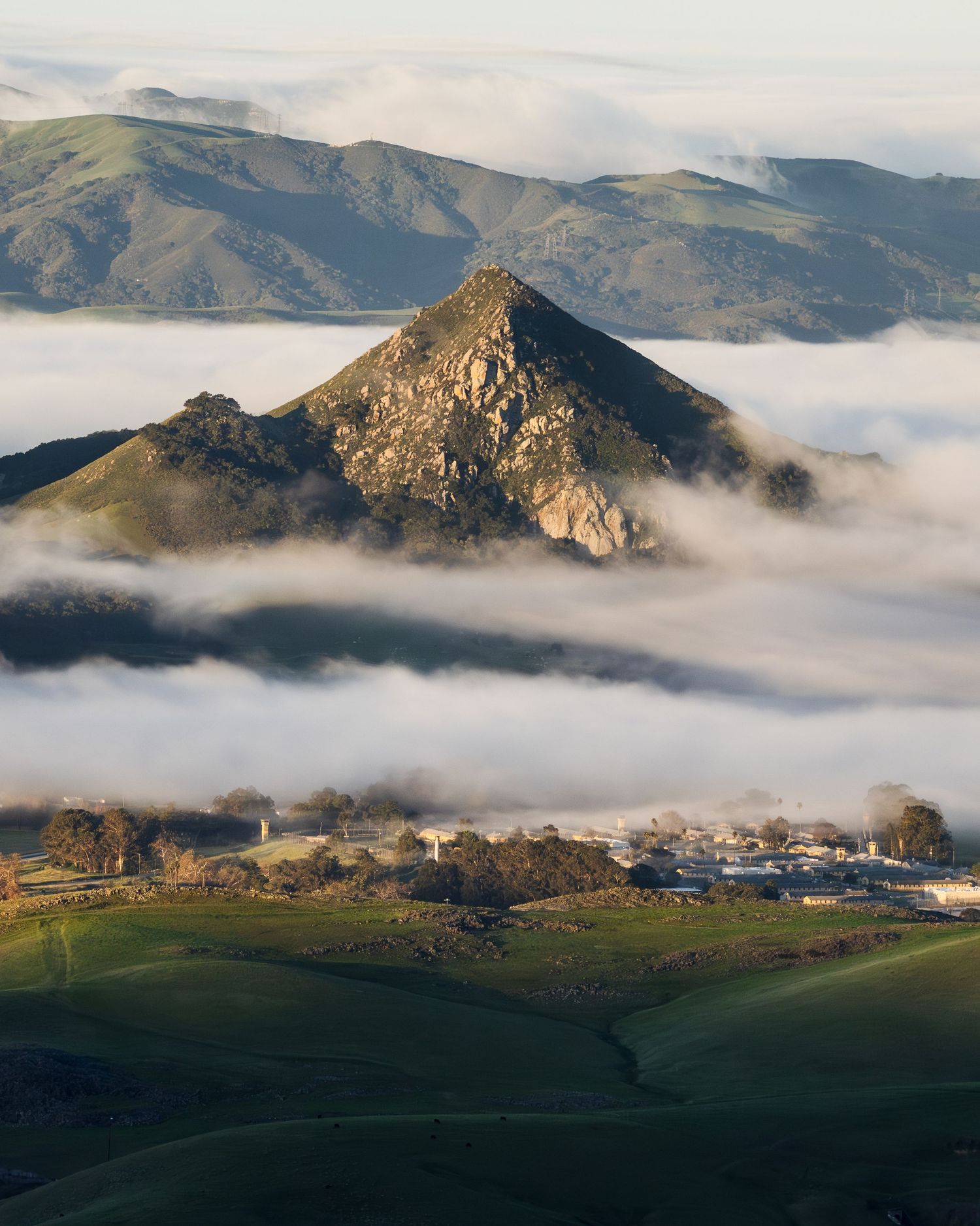 Pyramid-shaped rocky peak rising above a white cloud layer with sunlit green hills and mountains behind.